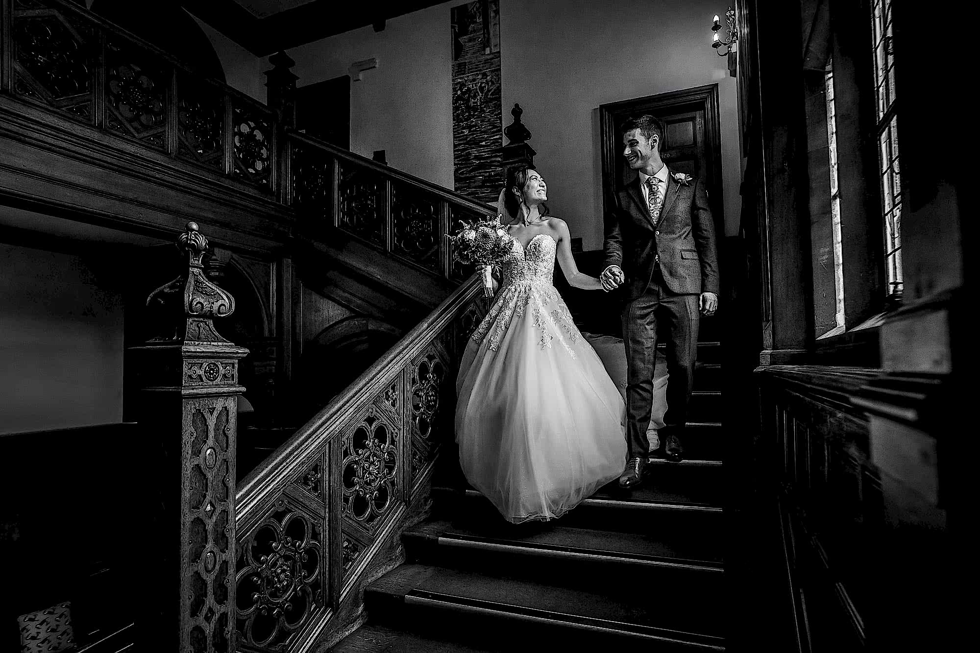 Black and white photo of wedding couple walking down the main staircase at Frensham Hall Wedding. Bride has a lovely flowing dress with a tight bodice and lace detail.