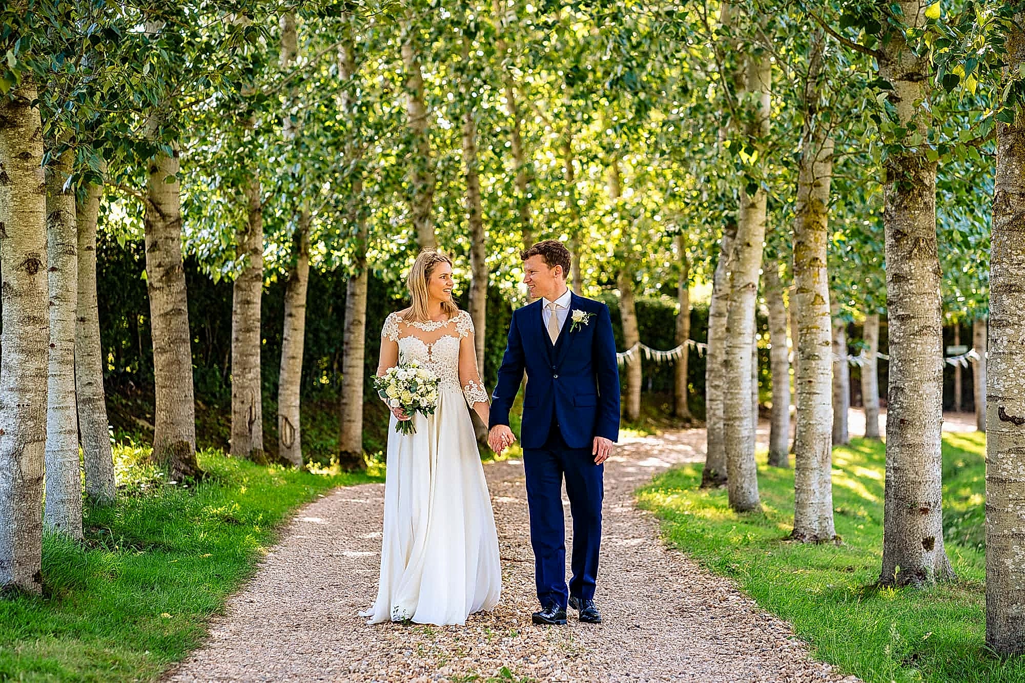 Wedding portrait of bride and groom walking along tree lined driveway, with filtered sunlight, captured at Rumbolds Farm wedding venue, by Paul at Tansley Photography